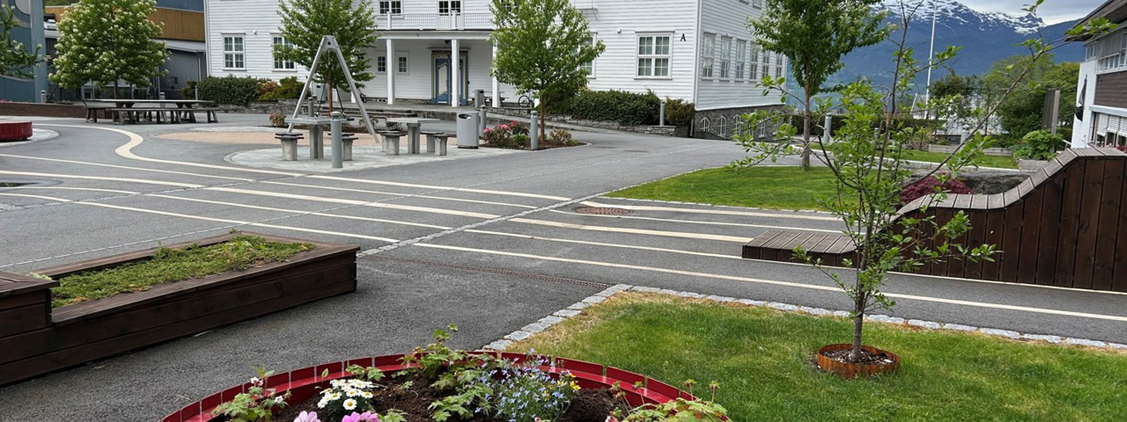Kvitt skulebygg med rødt tak og skuleplassen med blomsterkasse i framgrunnen.