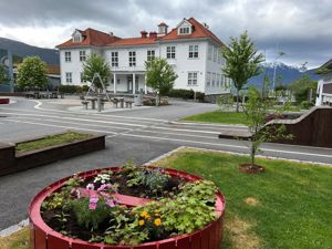 Kvitt skulebygg med rødt tak og skuleplassen med blomsterkasse i framgrunnen.