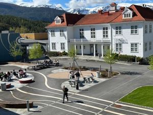 Kvitt trebygg med rødt tak og skuleplassen i framgrunnen.