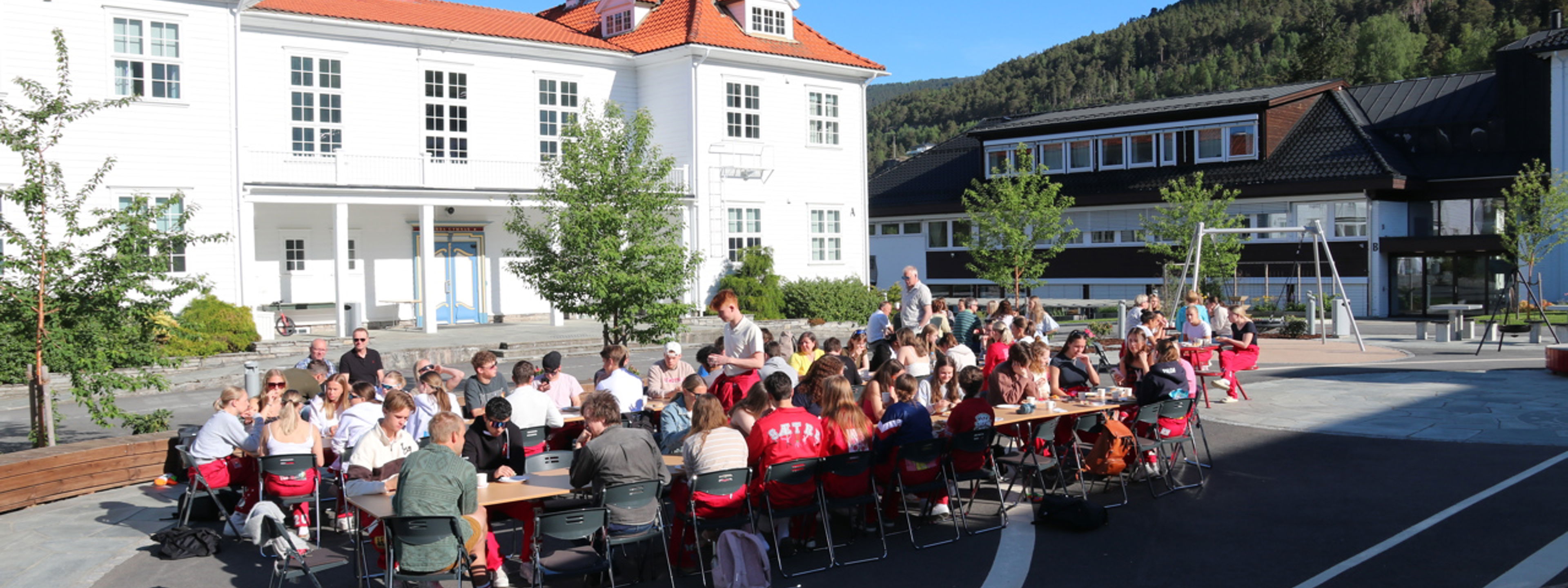 Russen sit rundt langbord på skuleplassen og et frukost.
