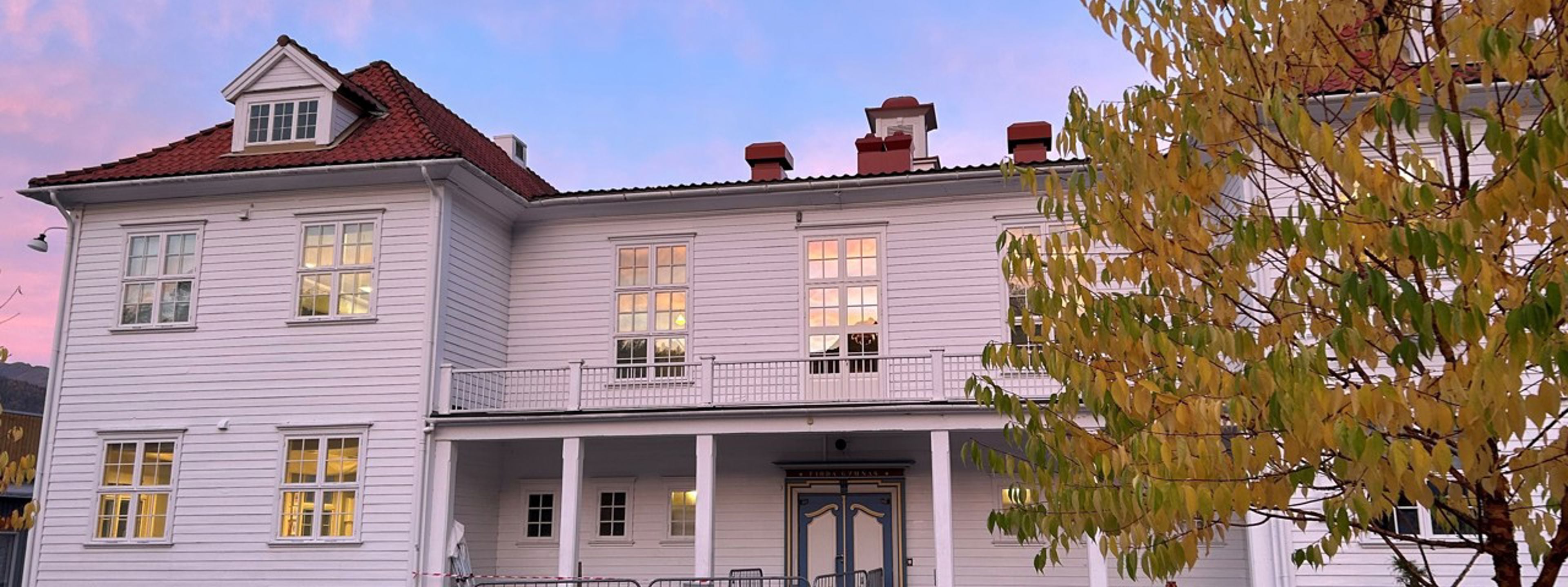Kvitt trebygg med blå himmel i bakgrunnen og eit haustleg tre med brune og oransje blad.